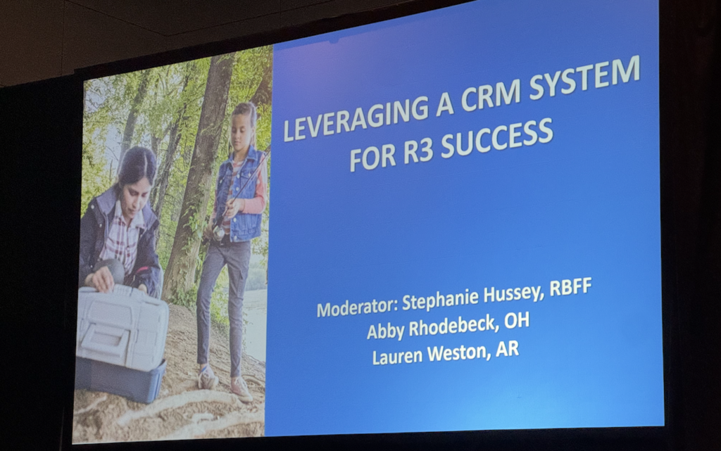 Conference presentation slide titled 'Leveraging a CRM System for R3 Success.' The slide features a blue background with white text listing the moderator, Stephanie Hussey from RBFF, and panelists Abby Rhodebeck (OH) and Lauren Weston (AR). On the left side of the slide, there is an image of two young girls outdoors near a body of water; one is kneeling and opening a tackle box, while the other stands holding a fishing rod.